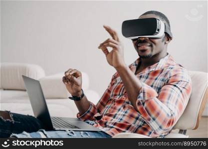 Distant work and futuristic gaming. African american man in vr goggles working with laptop at home. Relaxed freelancer clicks virtual buttons. Internet technology for business and entertainment.. Distant work and futuristic gaming. African american man in vr goggles working with laptop at home.