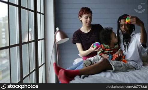 Diverse family with toddler son relaxing together in the bedroom at home. Cheerful interracial family spending great time together indoors. Smiling african father, joyful mother and curly mixed race child sitting on bed while playing with toys.