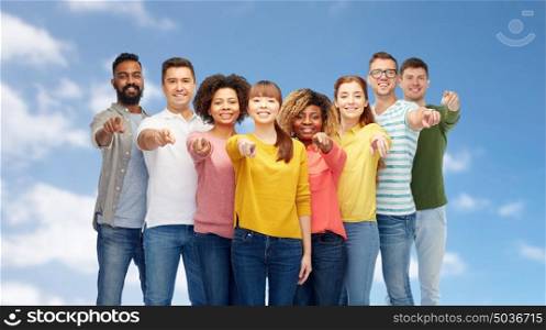 diversity, choice, ethnicity and people concept - international group of happy smiling men and women pointing finger on you over blue sky and clouds background. international group of people pointing on you
