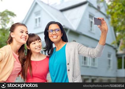 diversity, friendship and people concept - international group of happy smiling different women taking selfie with smartphone over house background. international group of happy women taking selfie