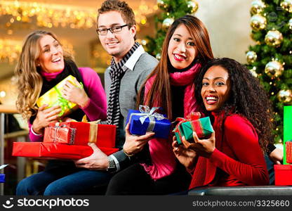 Diversity group of four people - Caucasian, black and Asian - sitting with Christmas presents and bags in a shopping mall in front of a Christmas tree with baubles