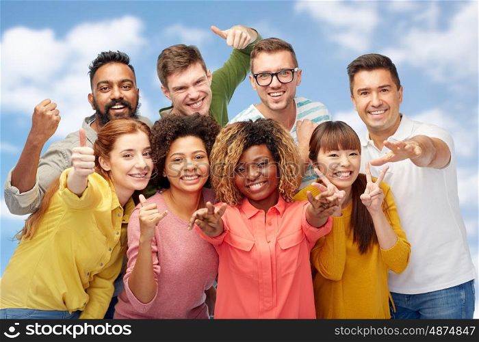 diversity, race, ethnicity and people concept - international group of happy smiling men and women showing thumbs up and peace over blue sky and clouds background