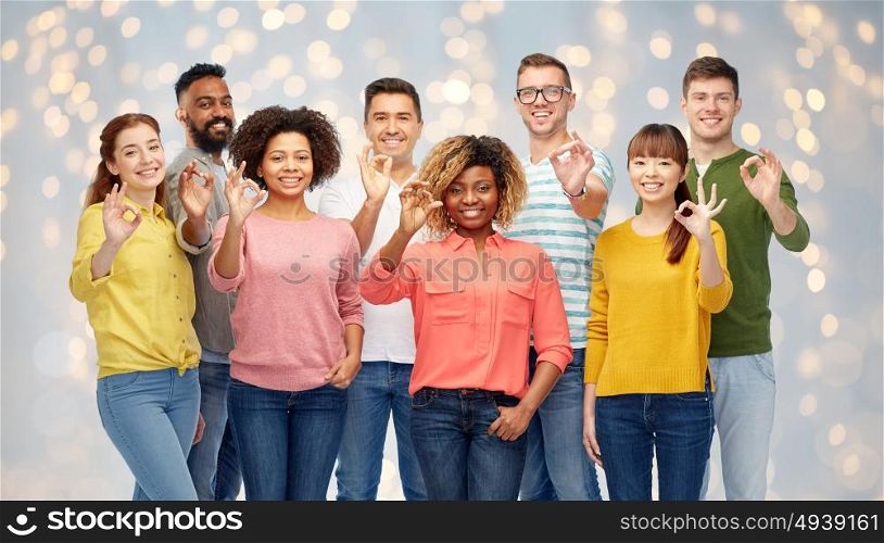 diversity, race, ethnicity and people concept - international group of happy smiling men and women showing ok hand sign over holidays lights background. international group of happy people showing ok