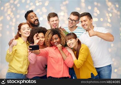 diversity, race, ethnicity, technology and people concept - international group of happy smiling men and women taking selfie by smartphone over holidays lights background