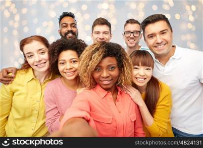 diversity, race, ethnicity, technology and people concept - international group of happy smiling men and women taking selfie over holidays lights background
