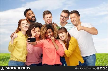 diversity, race, ethnicity, technology and people concept - international group of happy smiling men and women taking selfie by smartphone over blue sky and grass background
