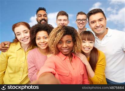diversity, race, ethnicity, technology and people concept - international group of happy smiling men and women taking selfie over blue sky and clouds background