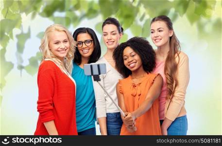 diversity, race, ethnicity, technology and people concept - international group of happy smiling different women taking picture with smartphone on selfie stick over green natural background. international group of happy women taking selfie