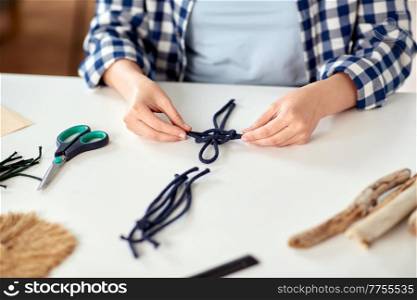 diy, handmade and hobby concept - close up of woman making macrame craft and knotting cords on table at home. woman making macrame and knotting cords
