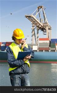 Docker in front of a huge containership being unloaded by a crane, using his cb radio, and reading his instructions from a clipboard
