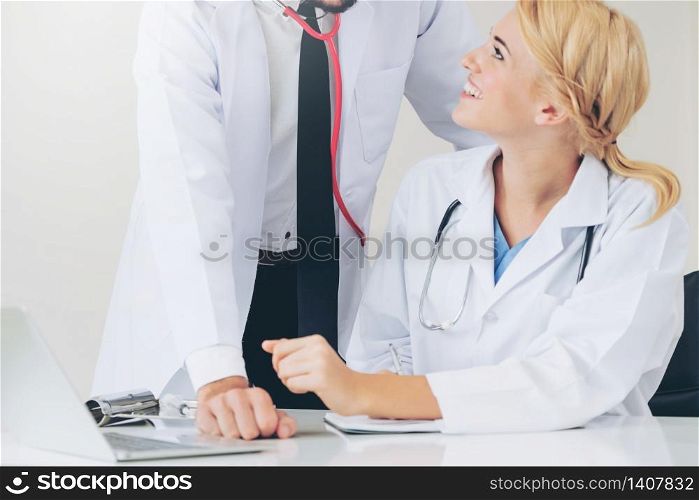 Doctor at hospital office writes notes on patients report while having conversation with another doctor that standing beside her.. Doctor writes report at desk with another doctor.