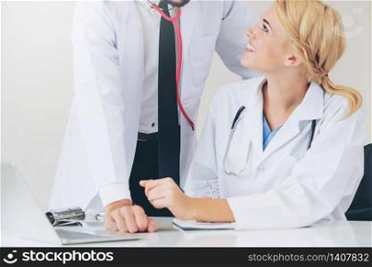 Doctor at hospital office writes notes on patients report while having conversation with another doctor that standing beside her.. Doctor writes report at desk with another doctor.