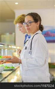 Doctor dressed with a coat and wearing a stethoscope serves herself healthy food at a cafeteria on an out of focus background. Healthy food concept.. Doctor serves herself food at a cafeteria