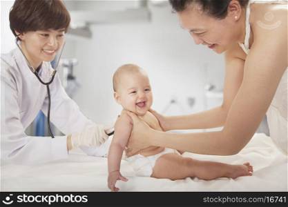 Doctor examining the baby with a stethoscope in the doctors office, mother holding the baby