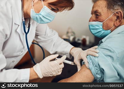 Doctor holding syringe with vaccine and making injection to senior patient with medical mask. Covid-19 or coronavirus vaccine. Physician wearing white coat and gloves using face mask