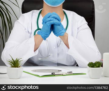 doctor in a white coat and blue latex gloves sits at a white work table in his office, concept of patient reception in the clinic, hands joined in front of the chest, focused pose