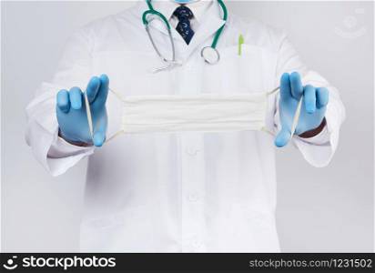 doctor in a white coat, blue latex sterile gloves holds textile medical masks in his hand on a white background, homemade mask made of white gauze and elastic