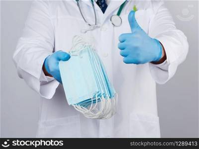 doctor in a white coat, blue latex sterile gloves holds textile medical masks in his hand on a white background, other hand shows like, protective accessory against viruses and bacteria