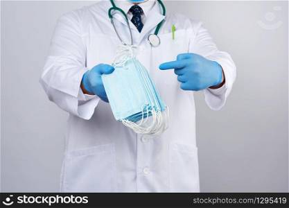 doctor in a white coat, blue latex sterile gloves holds textile medical masks in his hand on a white background, other hand shows like, protective accessory against viruses and bacteria