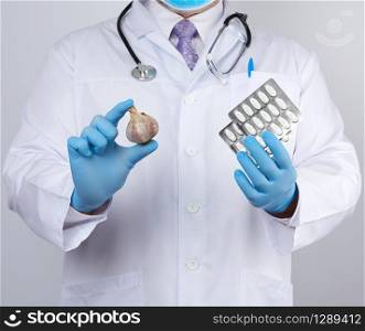 doctor in a white medical coat and blue rubber gloves holds a fresh head of garlic and pills, prevention against viral diseases, traditional medicine