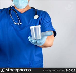 doctor in blue uniform and latex gloves is holding an empty plastic container for taking urine samples, gray background