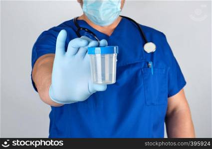 doctor in blue uniform and latex gloves is holding an empty plastic container for taking urine samples, close up