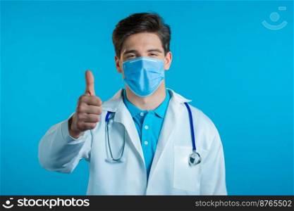 Doctor in mask showing thumb up sign over blue background. Positive young man in medical coat smiles to camera. Winner. Success. Body language. High quality photo. Doctor in mask showing thumb up sign over blue background. Positive young man in medical coat smiles to camera. Winner. Success. Body language.