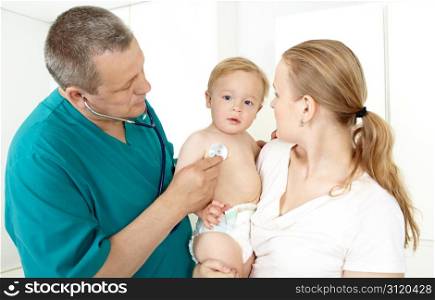Doctor, middlea??aged, in a green smock, is checking a baby with a stethoscope in a light medical study. The baby is 16 months old. Hea??s sitting on his muma??s laps. Caucasian, white.