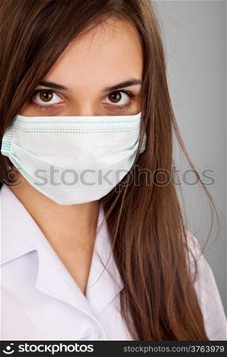 Doctor / nurse smiling behind surgeon mask. Closeup portrait of young caucasian woman model in white medical scrub isolated on a grey background