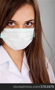 Doctor / nurse smiling behind surgeon mask. Closeup portrait of young caucasian woman model in white medical scrub isolated on a grey background