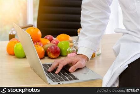 Doctor nutritionist in white lab coat at work, sitting on wooden desk and typing dietary consultation on a notebook computer keyboard. Healthcare and diet plan concept