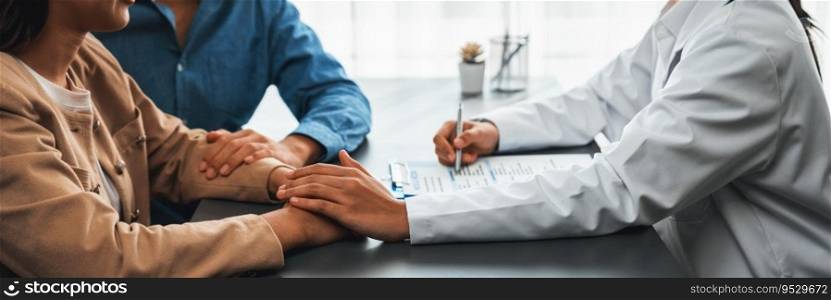 Doctor providing compassionate healthcare consultation while young couple patient holding hand, comfort each other after infertile report. Reproductive and medical fertility consulting. Neoteric. Infertile and medical consultation between young couple and doctor. Neoteric