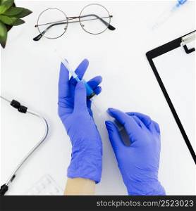 doctor s hand wearing blue gloves holding syringe white desk with stethoscope eyeglasses pills clipboard