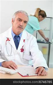 doctor sitting at desk with clipboard and nurse in background
