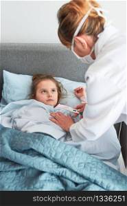 Doctor visiting little patient at home. Measuring the temperature of sick girl lying in bed. Woman wearing uniform and face mask. Medical treatment