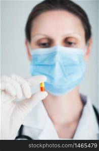 Doctor wearing blue face medical mask and clear latex gloves holding tablet pill on grey hospital wall background. Antibiotics and virus treatment tablet.