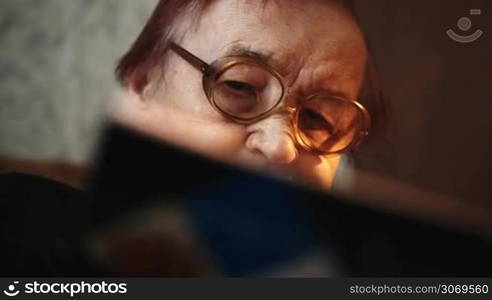 Dolly close-up shot of old woman in glasses reading a book at home