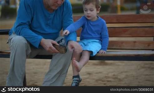Dolly shot of a boy and his grandpa on the wooden bench. Grandfather cleaning the shoe from the sand and helping the kid to put it on