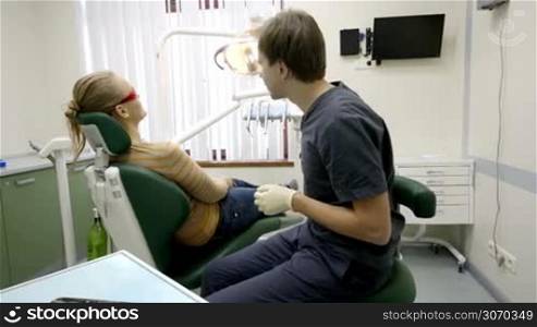 Dolly shot of a dentist starting examination of a patient. At first he fixing arm-chair and lamp and then taking tools