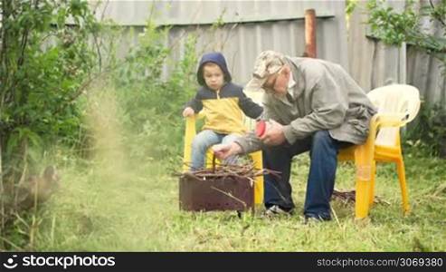 Dolly shot of a grandfather and a grandson sitting by the fire in the yard. Grandpa making a fire at first and then they watching it