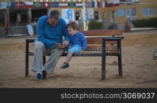 Dolly shot of a grandfather and a grandson sitting on the bench. Grandpa helping the little boy to put on the shoe