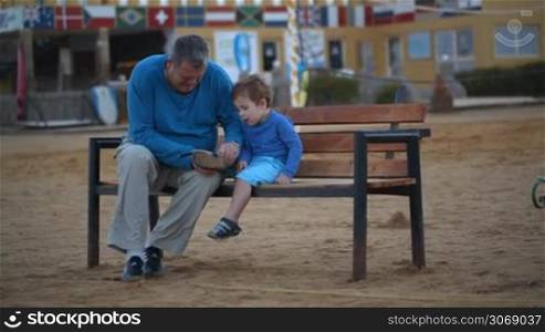 Dolly shot of a grandfather and a grandson sitting on the bench. Grandpa helping the little boy to put on the shoe