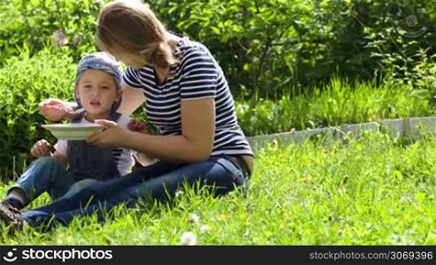 Dolly shot of a mother feeding her son sitting on the grass outdoor on a sunny summer day