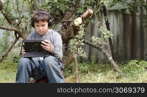 Dolly shot of a teenager boy sitting outdoor in headphones and using tablet computer