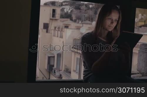 Dolly shot of a young smiling woman typing on touch pad. She sitting on the windowsill in the evening, city view behind