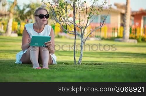 Dolly shot of beautiful woman wearing sunglasses sitting barefoot on a lawn in the shade of a tree with a laptop computer on her lap