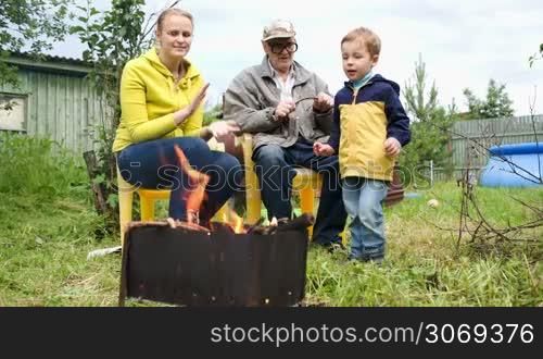 Dolly shot of mother, son and grandfather by the fire in the yard. Boy throwing firewood into the fire, mother applauding