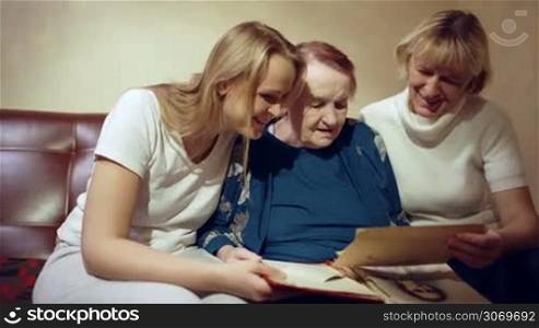 Dolly shot of three women of different generations looking through old family photobook and smiling