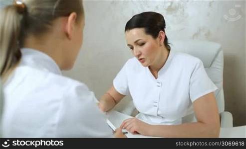 Dolly shot of two woman cosmeticians having talk about their job with pad on the table
