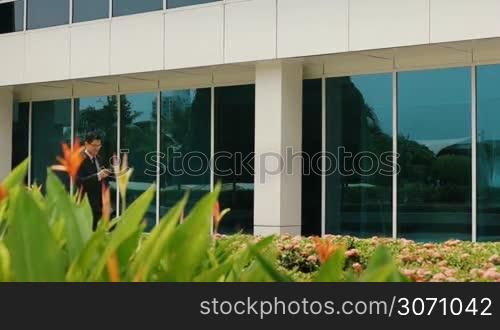 Dolly shot of young chinese businessman commuting to office with computer bag and reading message on mobile phone. The man smiles and walks out of frame